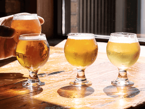 A beer tasting session with a hand raising a small glass of beer, among three other glasses on a wooden table, demonstrating the application of the EasyDens by Anton Paar Digital Density Meter for sampling and analyzing beer quality.