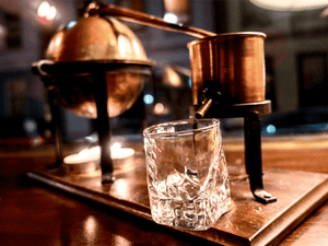 Copper distillery apparatus and a clear glass on a wooden table, potential application for EasyDens by Anton Paar Digital Density Meter in spirits' density measurement.